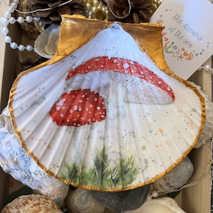 May include: A white seashell with a gold rim, painted with a red and white mushroom with white spots. The mushroom is surrounded by green grass and the text "She dreams of the sea" is written on a white card.