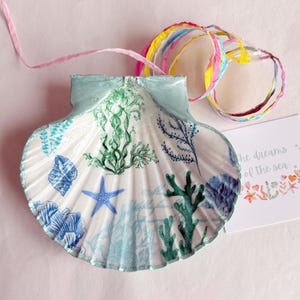 May include: A white scallop shell decorated with blue and green illustrations of seaweed, coral, and a starfish. The shell features a light blue painted edge and is attached to a multi-coloured ribbon. A small card reads the dreams of the sea.
