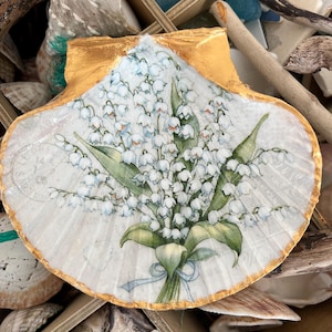 May include: A white seashell with a gold rim, painted with a bouquet of white lily of the valley flowers with green leaves. The shell is sitting in a wooden box with other seashells.