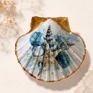 May include: A decorative scallop shell featuring a printed design of various seashells and a starfish in shades of blue and white. The edges and the top hinge of the shell are painted with a metallic gold finish, creating a distinct border.