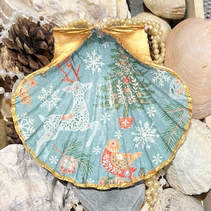 May include: A blue and gold seashell dish with a Christmas-themed design. The design features a deer, a Christmas tree, snowflakes, and a bird. The seashell is trimmed with gold paint.