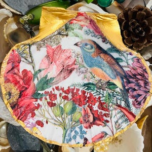 May include: A white seashell with a gold rim, decorated with a colourful floral and bird design. The shell is filled with a variety of flowers, including red poppies, pink lilies, and yellow daisies. A blue bird with orange accents is perched on a branch in the centre of the shell.
