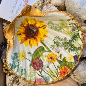 May include: A large, white seashell with a gold-painted rim, decorated with a vibrant floral design featuring a sunflower and other colourful flowers. The shell is surrounded by other shells and jewellery.