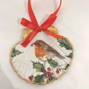May include: A white seashell ornament with a gold trim, featuring a robin perched on a holly branch with red berries. The ornament has a red ribbon for hanging.
