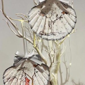 May include: Two white seashell ornaments with a silver trim. Each ornament features a hand-painted illustration of a white rabbit with a red robin bird. One ornament shows the rabbit holding a sprig of holly, while the other shows the rabbit holding a red stocking.