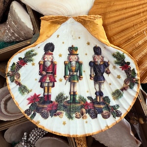 May include: A large, decorated seashell featuring three toy nutcracker soldiers. The shell is painted with a white background, gold trim, and festive holiday decorations. The nutcrackers are in red, green, and blue uniforms.