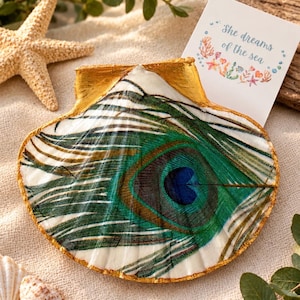 May include: A scallop shell painted with a vibrant green and blue peacock feather pattern, featuring a metallic gold painted edge. A small white card is tucked behind the shell, displaying the text She dreams of the sea with colorful illustrations.