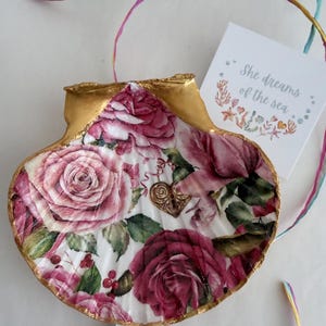 May include: A decorated seashell with a gold rim and a floral rose pattern. The shell has a heart-shaped charm and a tag that reads "She dreams of the sea."