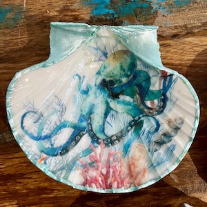 May include: A painted scallop shell with an octopus design. The octopus is in blue and green hues, with coral and seaweed details. The shell has a light blue border and is set against a wooden background.