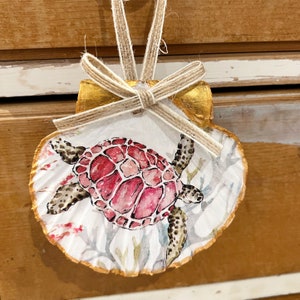 May include: A hand-painted seashell ornament featuring a pink and white sea turtle with black accents. The seashell is painted gold around the edges and has a white background with a watercolour design of coral and seaweed.