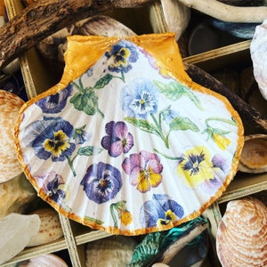 May include: A large, white scallop shell decorated with a floral pattern of purple, yellow, and blue pansies. The shell's edge is painted gold, and the background includes other shells and driftwood.