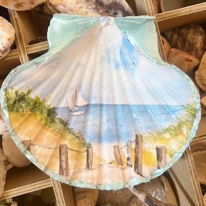 May include: A scallop shell featuring a hand-painted coastal scene with a sailboat on blue water, sandy dunes with green grass, and wooden fence posts. The shell has a light blue painted border and a textured, ribbed surface with natural ridges.