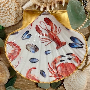 May include: A white seashell dish with a gold rim, featuring a painted design of a lobster, crab, and mussels. The dish is ideal for holding small items such as jewellery or trinkets.