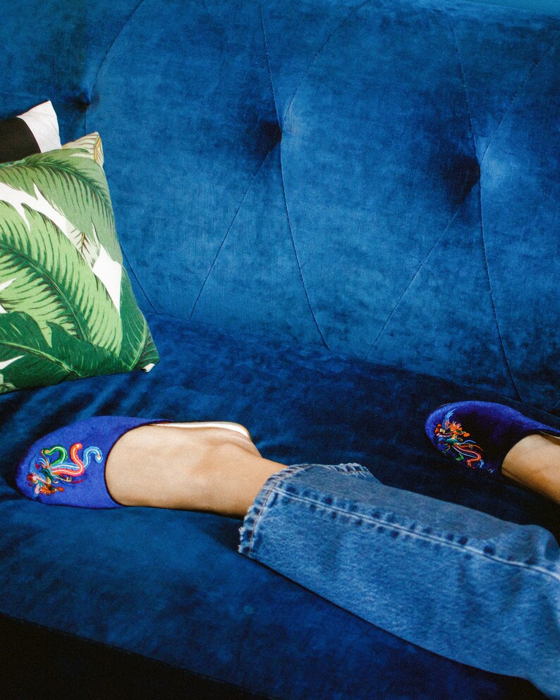 May include: A pair of blue velvet slippers with colorful embroidery on a blue velvet couch. The slippers are worn by a person wearing blue jeans.