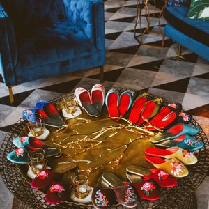 May include: A collection of embroidered velvet slippers in various colors and patterns, displayed on a gold tray with a decorative branch design. The slippers are arranged in a circle, with the toes pointing outwards.