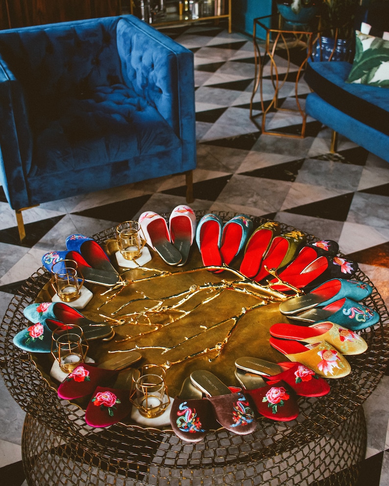 May include: A collection of embroidered velvet slippers in various colors and patterns, displayed on a gold tray with a decorative branch design. The slippers are arranged in a circle, with the toes pointing outwards.