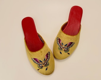 May include: A pair of gold velvet slippers with red soles and embroidered butterfly designs. The butterflies have colorful wings with purple, blue, green, and yellow accents.