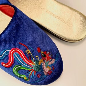 May include: A pair of blue velvet slippers with a colorful embroidered dragon design. The slippers have a gold-colored sole.