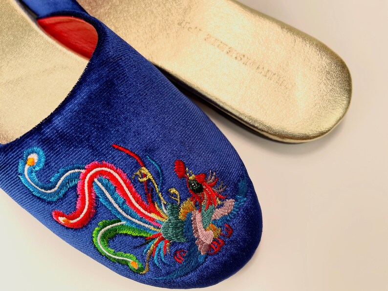 May include: A pair of blue velvet slippers with a colorful embroidered dragon design. The slippers have a gold-colored sole.