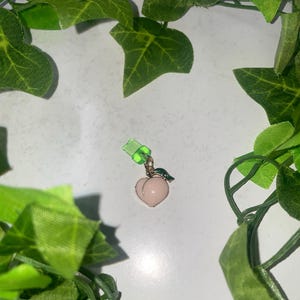 May include: A peach-shaped charm with a pink body, gold outline, and a small green leaf. The charm is attached to a green clasp. The image is framed by green ivy.