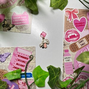 May include: A pink and white mushroom charm with a pink stem and a white cap. The charm is surrounded by green leaves and pink and white stickers with text such as "Crying Over Fictional Characters", "Romance Books All Day Everyday", "Shadow Daddies", "Fairy Smut", and "Tamlin's Tears".