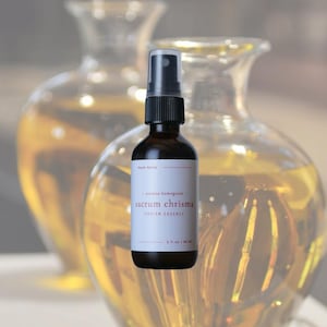 May include: A dark brown glass bottle with a black spray nozzle. The label reads "Sacrum Chrisma, Chrism Essence" and "2 fl oz / 60 ml". The background features two blurred glass vases filled with a golden liquid.