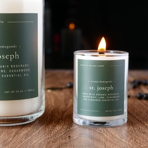 Op de afbeelding: Twee glazen potkaarsen op een houten oppervlak. De grotere kaars heeft een donkergroen etiket met witte tekst "Joseph". De kleinere kaars is aangestoken, met een vlam, en heeft een etiket "St. Joseph". De kleinere kaars is 85 g.