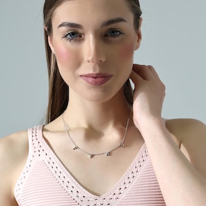 Può includere: Una donna indossa una canotta rosa chiaro con un delicato bordo a uncinetto. Indossa una collana d'argento con piccoli ciondoli.