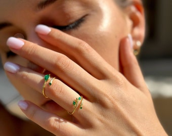 May include: A gold ring with a delicate, twisting design featuring small emerald green gemstones. The ring is worn on a finger.