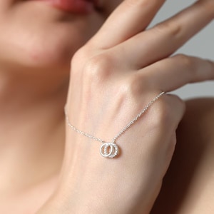 Può includere: Una delicata collana d'argento con due cerchi intrecciati, uno impreziosito da piccole gemme scintillanti. La collana è mostrata sulla mano di una persona, evidenziando il suo design elegante e i dettagli raffinati. Un accessorio perfetto.