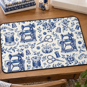 May include: A rectangular sewing mat featuring a repeating blue and white pattern of vintage sewing machines, spools of thread, scissors, thimbles, buttons, and floral motifs on a cream background with a black border.