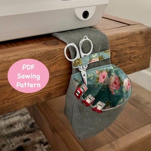 May include: A gray fabric sewing organizer with a floral interior, holding scissors and red clips, attached to a wooden table. The text "PDF Sewing Pattern" is on a pink circle.