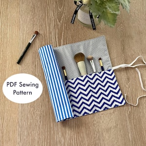 Op de afbeelding: Een blauwe en witte gestreepte stoffen make-up kwastrol met een zigzagpatroon aan de binnenkant. De rol heeft een witte lintband en is opgerold met kwasten erin.