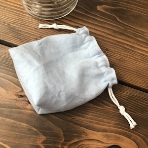 May include: A light blue linen drawstring bag with white cords. The bag is sitting on a wooden surface.