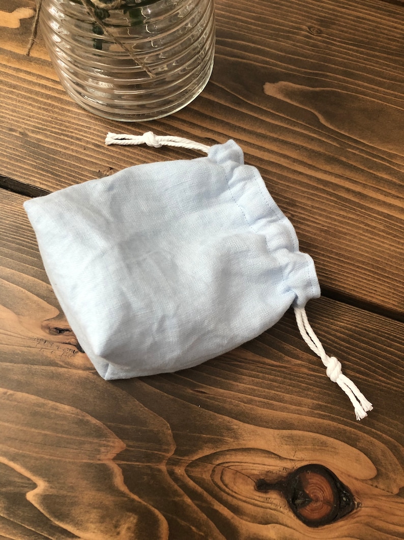 May include: A light blue linen drawstring bag with white cords. The bag is sitting on a wooden surface.