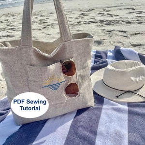 Op de afbeelding: Een beige linnen tas met een groot vak aan de voorkant. Het vak heeft een eenvoudig geborduurd ontwerp van een zonsondergang boven de oceaan. De tas ligt op een blauw-wit gestreepte strandhanddoek met een strohoed en een zonnebril.