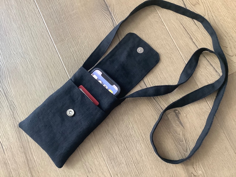 May include: A black fabric crossbody phone pouch with a long shoulder strap and a magnetic snap closure. The pouch features an open exterior pocket containing a smartphone and a red card, with the main compartment flap open to reveal the snap.