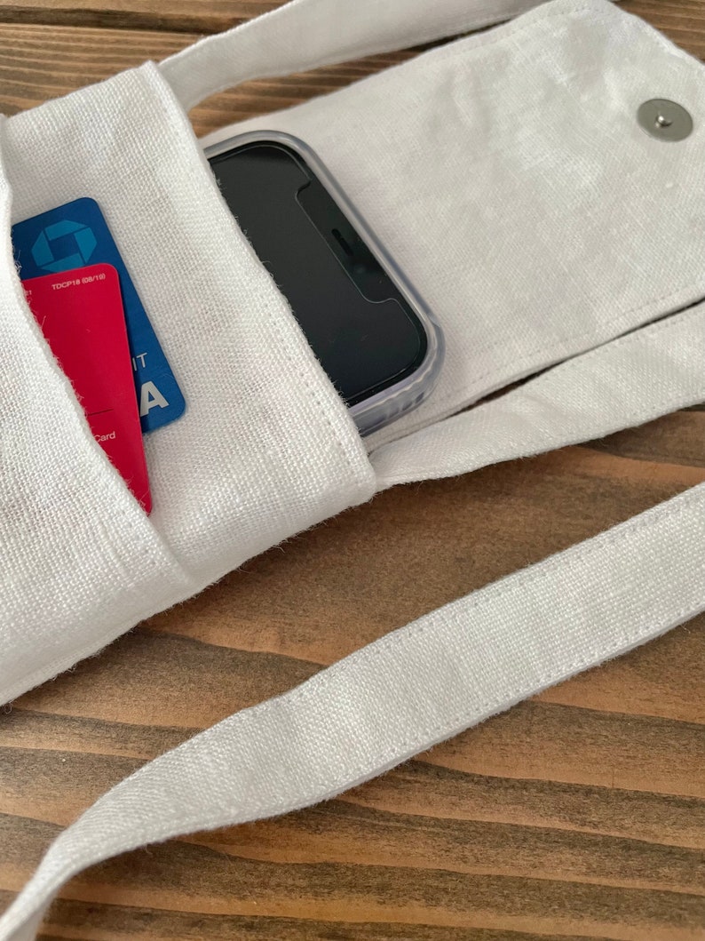 May include: A white fabric crossbody phone pouch with a flap closure secured by a silver metal snap. The pouch features an exterior pocket containing a blue credit card and a red credit card, while a black smartphone is tucked inside the main compartment.