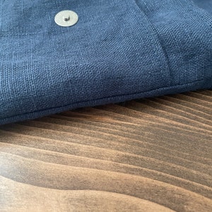 May include: A dark blue textile bag featuring a visible silver-toned circular magnetic snap closure. The fabric displays a distinct woven texture, and the bag is constructed with a clean, straight stitched seam along the top edge of the flap.