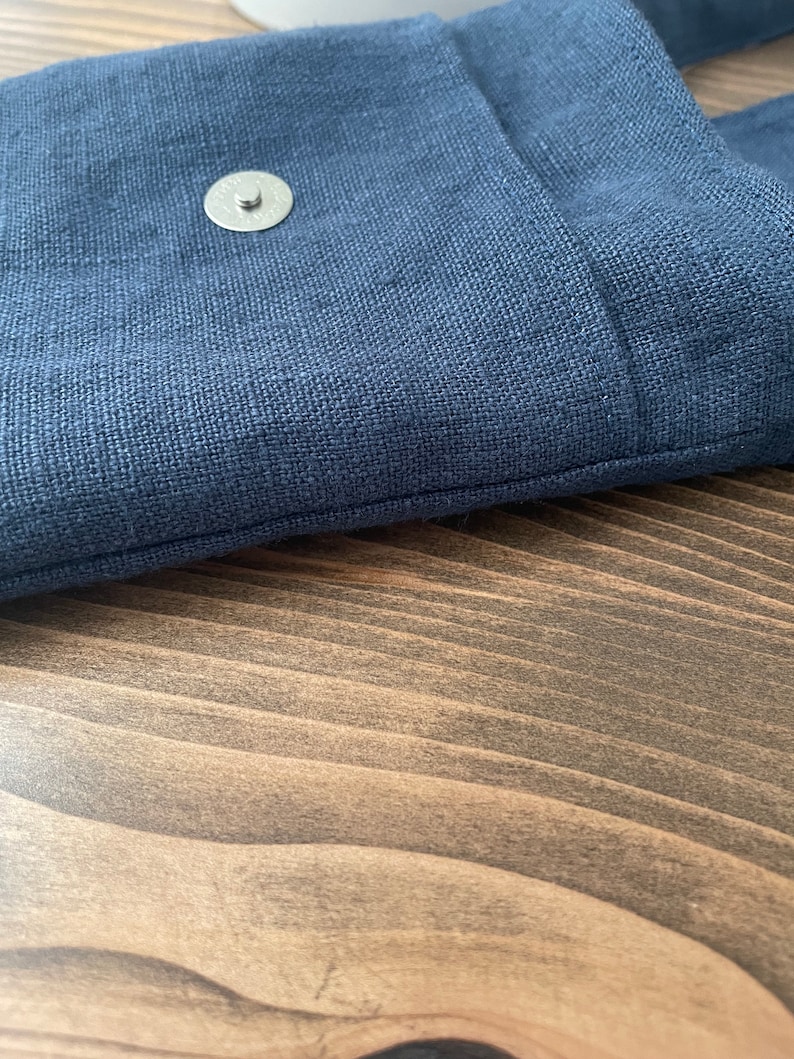 May include: A dark blue textile bag featuring a visible silver-toned circular magnetic snap closure. The fabric displays a distinct woven texture, and the bag is constructed with a clean, straight stitched seam along the top edge of the flap.