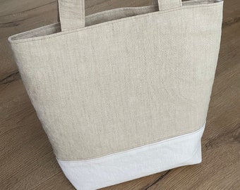 Könnte beinhalten: Eine beige Leinen-Tragetasche mit weißem Besatz. Die Tasche hat zwei lange Henkel.