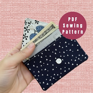 Può includere: Un portafoglio in tessuto fatto a mano con sfondo blu navy e motivo a triangoli bianchi. Il portafoglio è aperto, rivelando contanti e una carta di credito. L'interno presenta un motivo floreale. Il testo "PDF Sewing Pattern" è in un cerchio rosso.