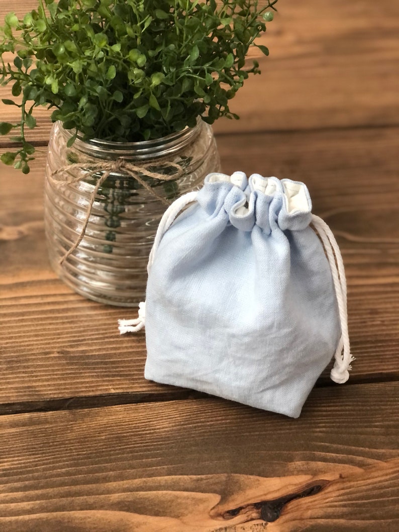 May include: A light blue linen drawstring bag with white lining and white drawstrings. The bag is sitting on a wooden surface.