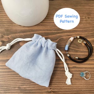 Könnte beinhalten: Eine hellblaue Leinentasche mit weißen Kordeln, ein schwarz-weißes Armband mit einem blau-weißen Anhänger und ein türkisfarbener Ring auf einer Holzoberfläche. Der Text "PDF Sewing Pattern" ist in einem weißen Kreis auf der Holzoberfläche geschrieben.