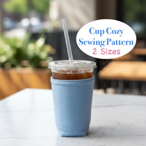 May include: A light blue cup cozy made of fabric, wrapped around a clear plastic cup filled with a beverage and a straw. The image includes text that reads "Cup Cozy Sewing Pattern 2 Sizes". The cup is on a white table.