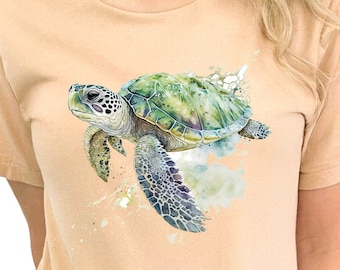 Puede incluir: Una camiseta marrón claro con una ilustración de acuarela de una tortuga marina verde nadando. La tortuga tiene un caparazón y aletas detalladas.