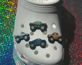 May include: White clog shoe with four monster truck shoe charms. The charms are green, blue, yellow, and black.