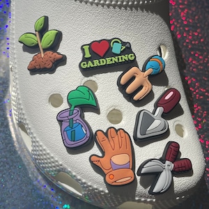 May include: A set of colorful gardening-themed shoe charms. The charms include a green plant, a red heart with the words "I love gardening", a watering can, a pair of gardening gloves, a trowel, a pair of pruning shears, and a plant in a glass jar.