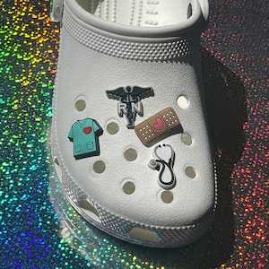 May include: White clogs with a variety of medical themed shoe charms. The charms include a stethoscope, a bandage with a heart, a scrub top with a heart, and a caduceus with the letters "RN".