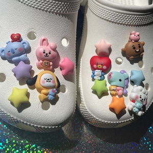 May include: White clogs with colorful cartoon character charms. The charms include a pink bunny, a brown bear, a yellow dog, a purple horse, a red heart, and several stars.